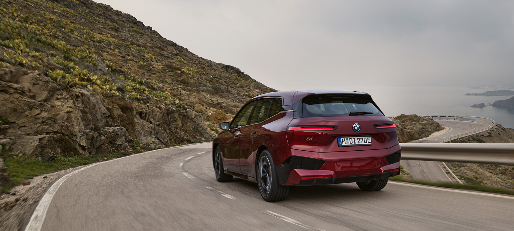 BMW iX i20 2021 SUV electric BMW iX xDrive50 Roşu Aventurin Imagine din partea din spate în timp ce se deplasează pe o şosea de coastă